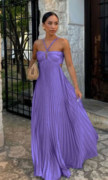Purple pleated maxi dress with crisscross straps, worn outdoors near stone wall and gate